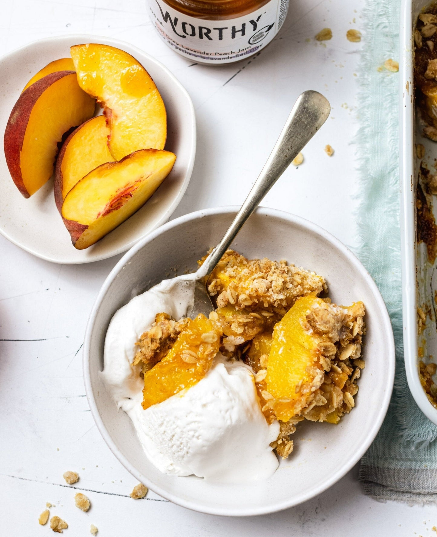 A tray of baked peach crisp with Worthy’s Earl Grey Lavender jam filling next to a portion of the crisp in a bowl with vanilla ice cream.