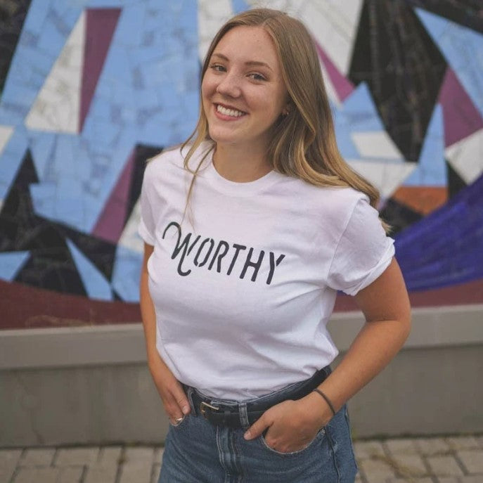 A woman smiling into the camera wearing a white unisex t-shirt with a black Worthy logo across the front while standing in front of a colourful background.