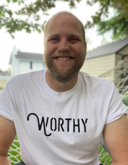 A man smiling into the camera wearing a white unisex t-shirt with a black Worthy logo across the front.