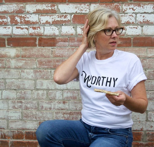 A woman looking to the left wearing a white unisex t-shirt with a black Worthy logo across the front and holding a piece of toast slathered in Worthy jam.