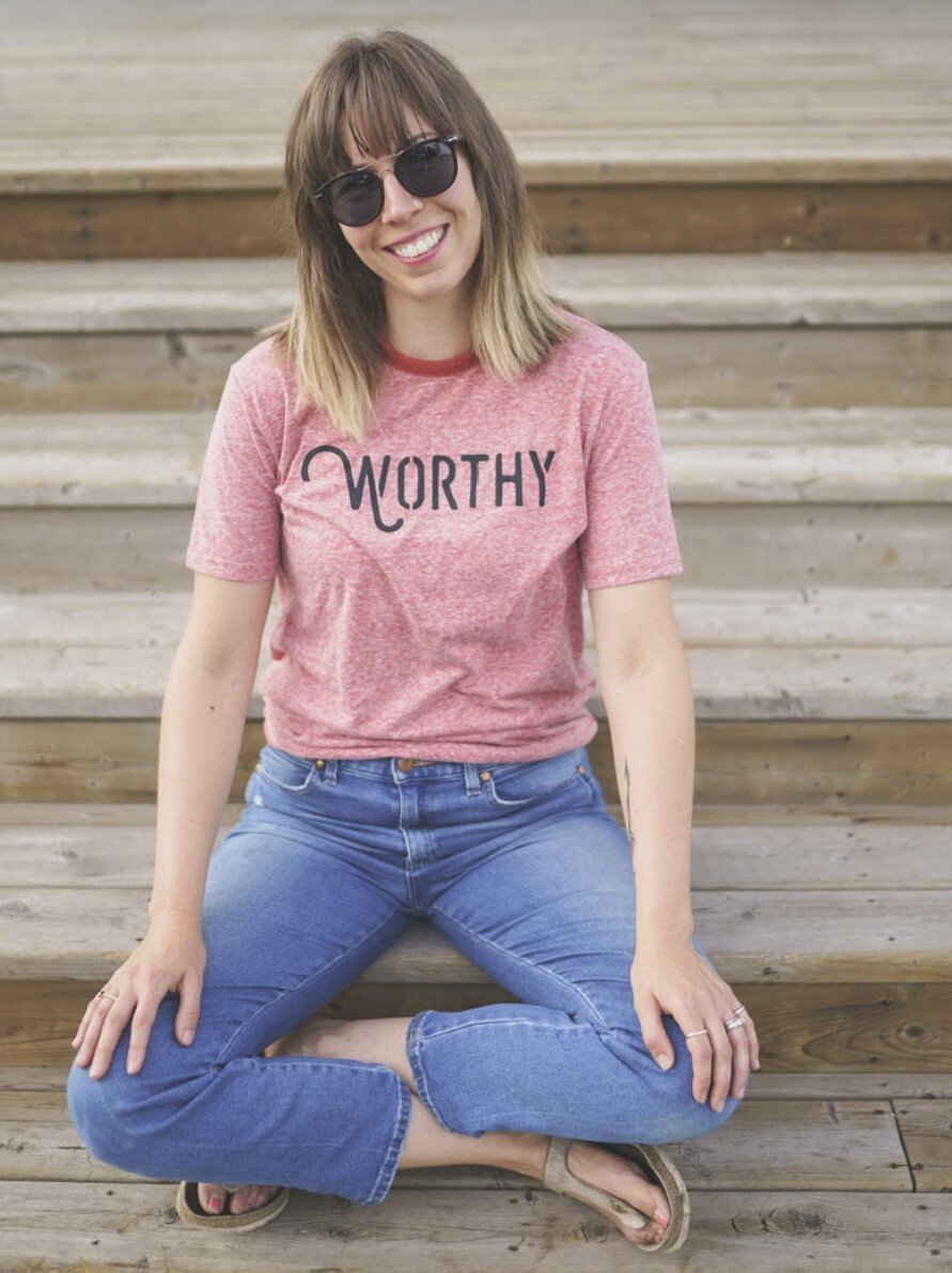 Ashlee, the founder of Worthy Jams, sitting crossed legged on wooden stairs wearing a pink worthy logo t-shirt