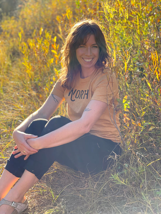 Ashlee, the founder of Worthy Jams, sitting in field during sunset wearing a gold worthy logo t-shirt