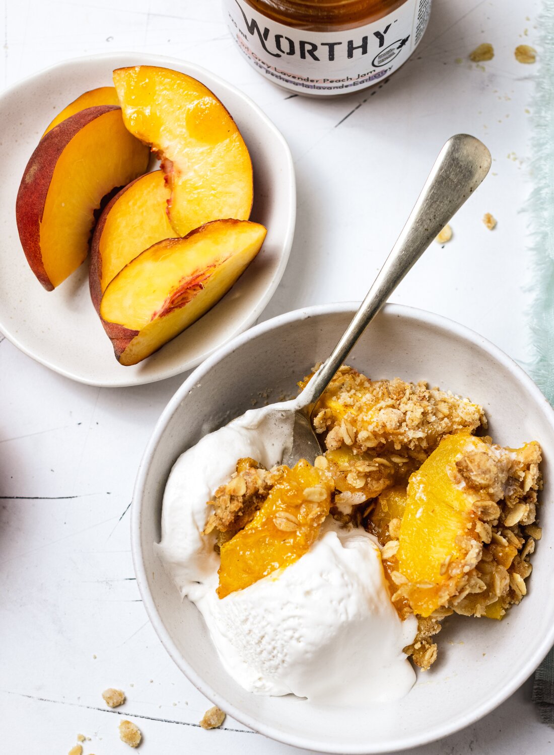 Baked peach crisp with Worthy’s Earl Grey Lavender jam filling in a bowl with vanilla ice cream next to a plate of peach slices.