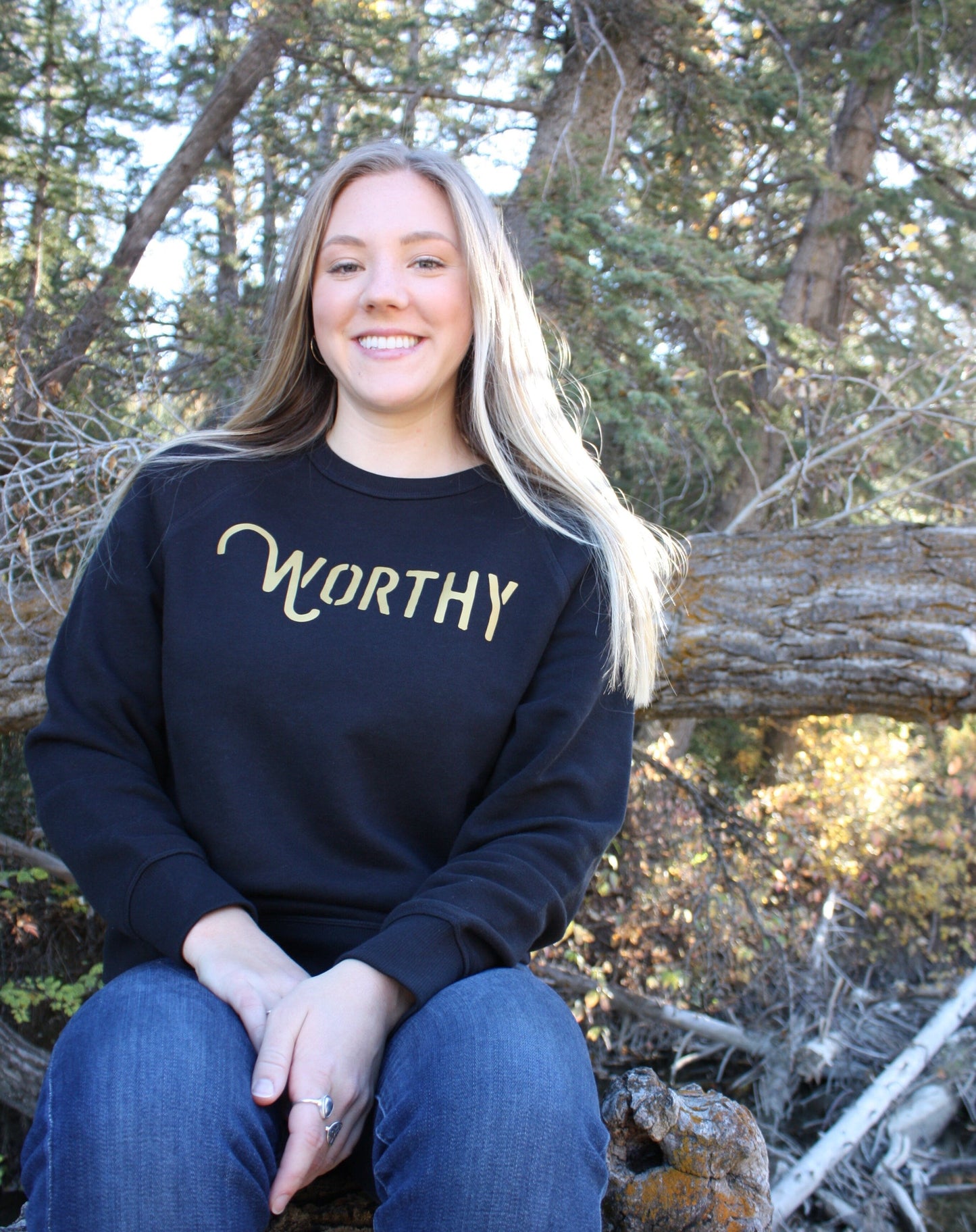 Woman sitting outside, wearing a fitted black pullover sweater with "WORTHY" printed across the front in gold lettering.