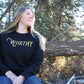 Woman sitting outside, wearing a fitted black pullover sweater with "WORTHY" printed across the front in gold lettering.