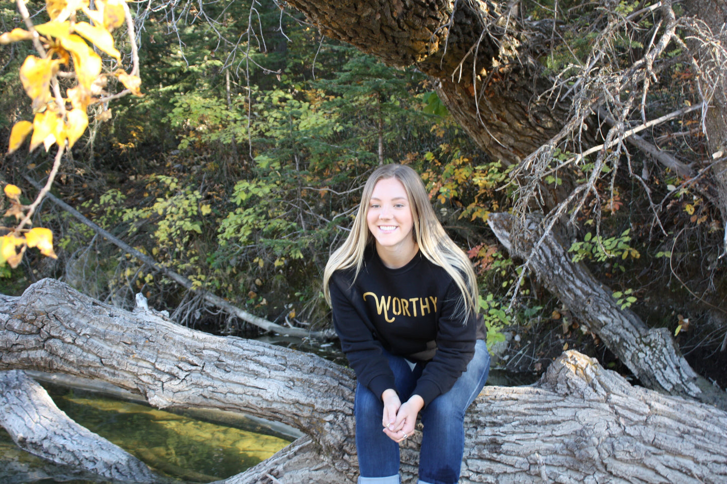 Woman sitting outside on a log, wearing a fitted black pullover sweater with "WORTHY" printed across the front in gold lettering.