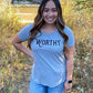 A woman smiling into the camera while standing on a hiking trail wearing a grey t-shirt with black "WORTHY" lettering across the front.