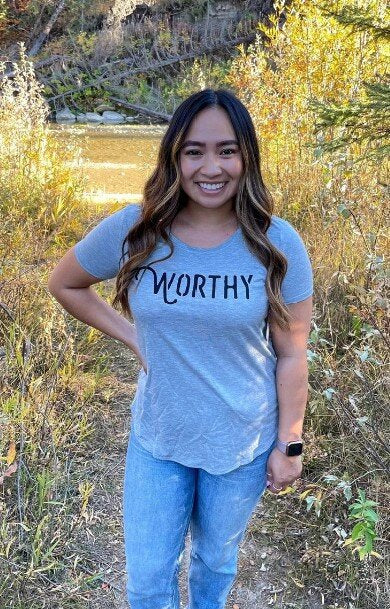 A woman smiling into the camera while standing on a hiking trail wearing a grey t-shirt with black "WORTHY" lettering across the front.