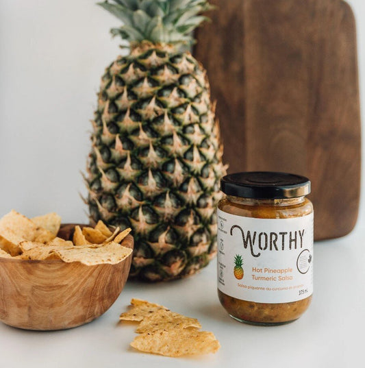 Worthy's Hot Pineapple Turmeric Salsa beside a wooden bowl of tortilla chips and a pineapple and cutting board in the background.