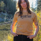 Woman standing beside a creek wearing a heathered yellow-gold tshirt with black "WORTHY" lettering across the front.