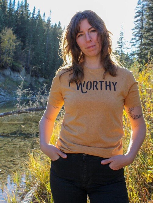 Woman standing beside a creek wearing a heathered yellow-gold tshirt with black "WORTHY" lettering across the front.