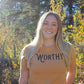 Woman smiling into the camera while standing outside wearing a heathered yellow-gold tshirt with black "WORTHY" lettering across the front.