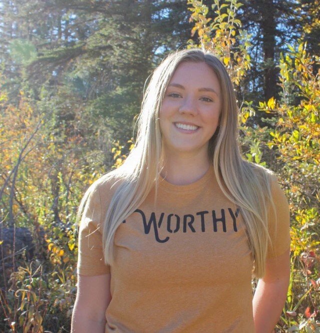 Woman smiling into the camera while standing outside wearing a heathered yellow-gold tshirt with black "WORTHY" lettering across the front.