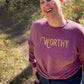 A woman standing in nature and laughing while wearing a dark pink heathered sweater with gold "WORTHY" lettering across the front.