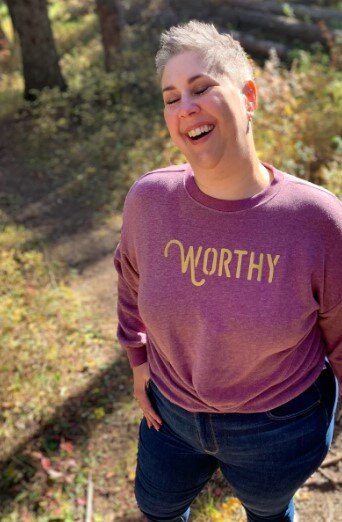 A woman standing in nature and laughing while wearing a dark pink heathered sweater with gold "WORTHY" lettering across the front.