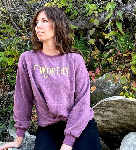 A woman sitting on a large rock staring off into the distance while wearing a dark pink heathered sweater with gold "WORTHY" lettering across the front.