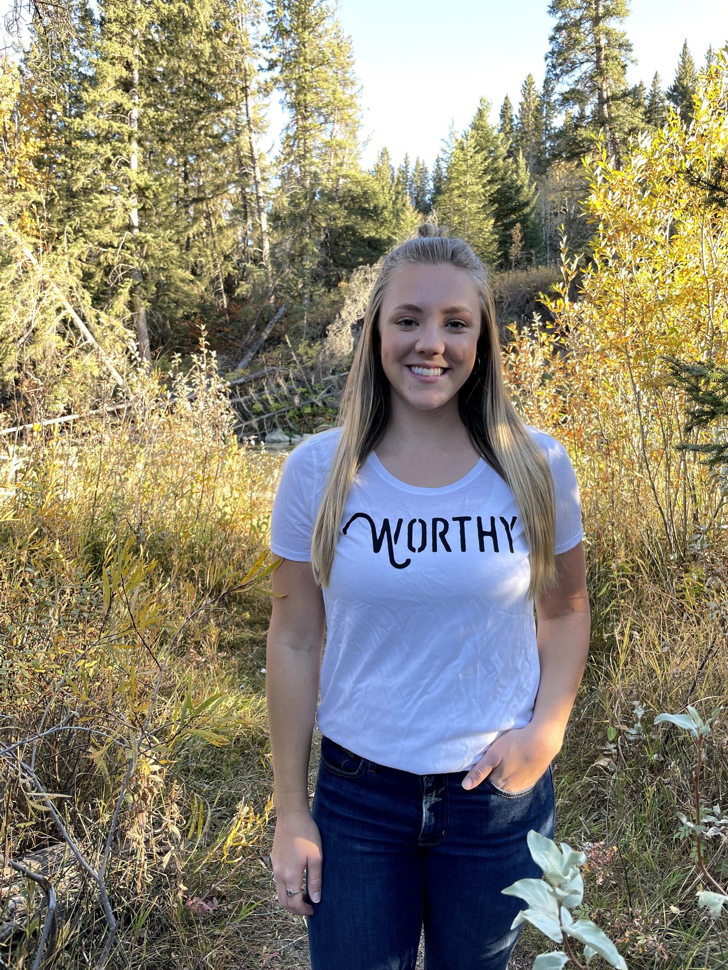 A woman standing in nature, wearing a white t-shirt with black "WORTHY" lettering across the front.