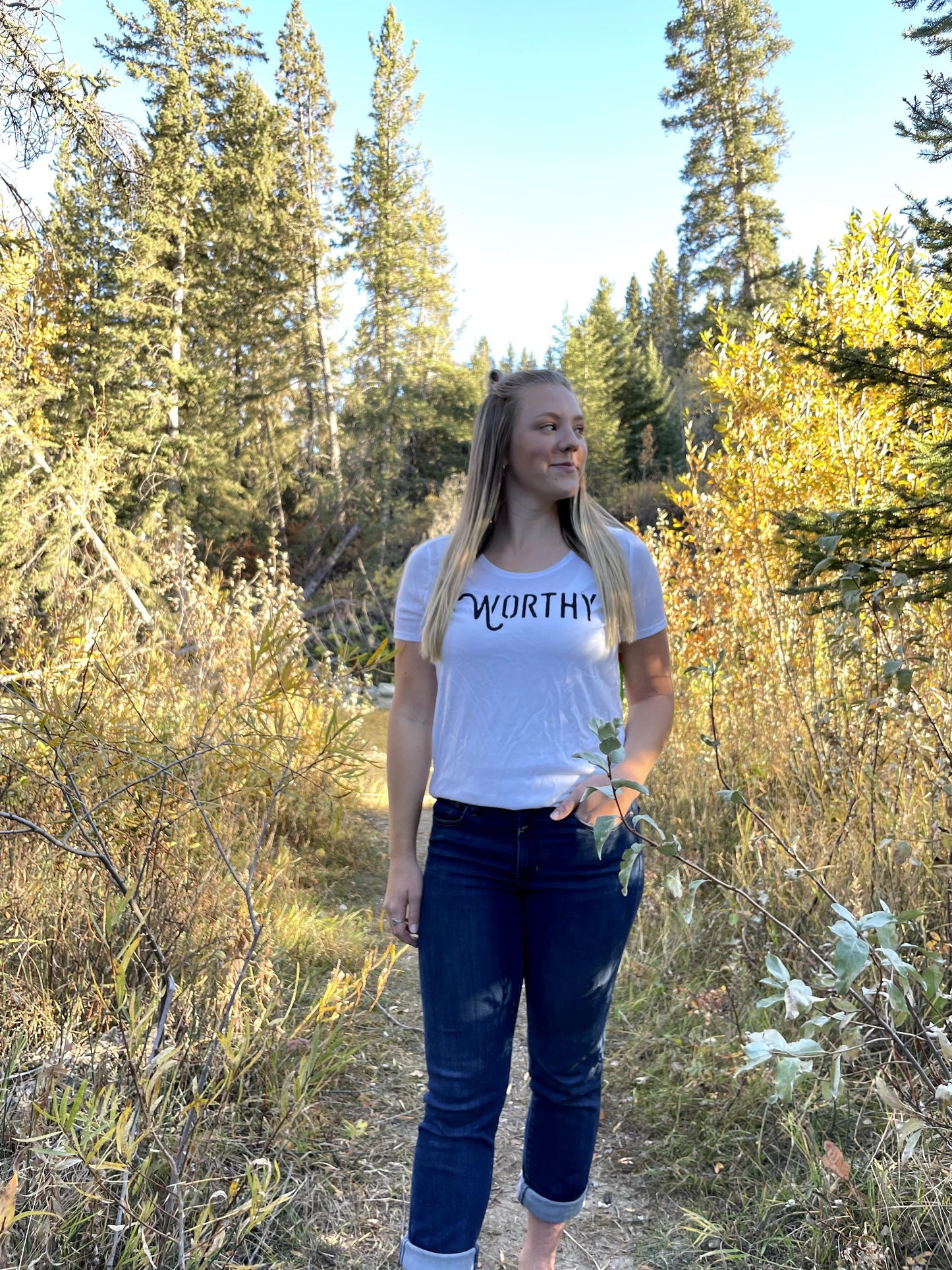 A woman standing in nature looking to the right while wearing a white t-shirt with black "WORTHY" lettering across the front.
