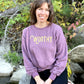 A woman sitting on a rock smiling into the camera wearing a dark pink heathered sweater with gold "WORTHY" lettering across the front.