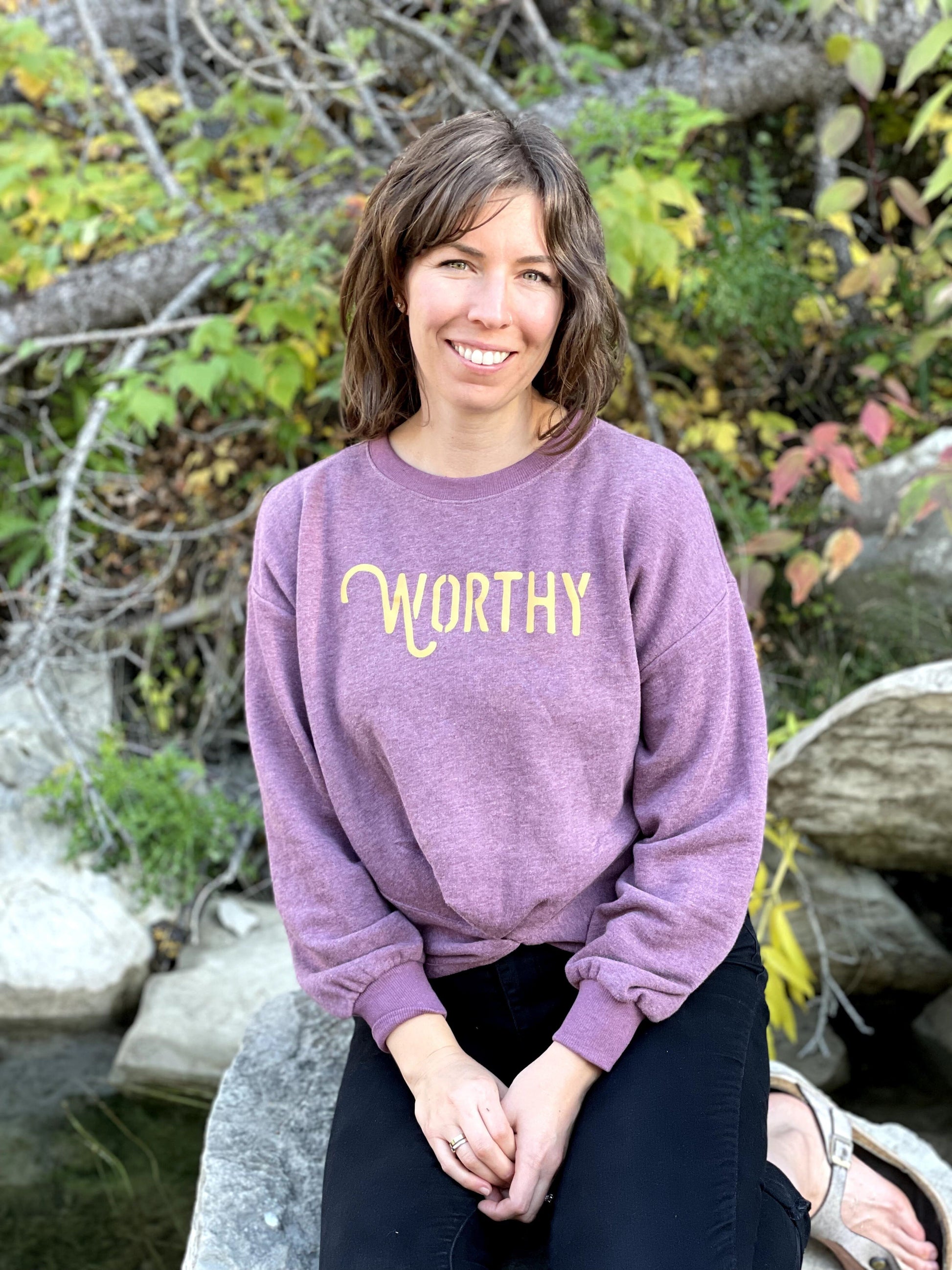 A woman sitting on a rock smiling into the camera wearing a dark pink heathered sweater with gold "WORTHY" lettering across the front.